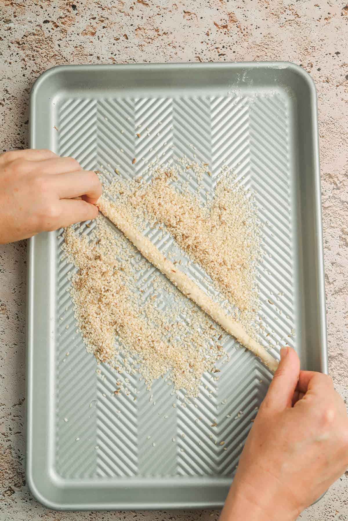 Two hands roll a long piece of dough in a tray filled with sesame seeds. The tray is metallic with ridged lines, and the surface beneath is speckled light brown.