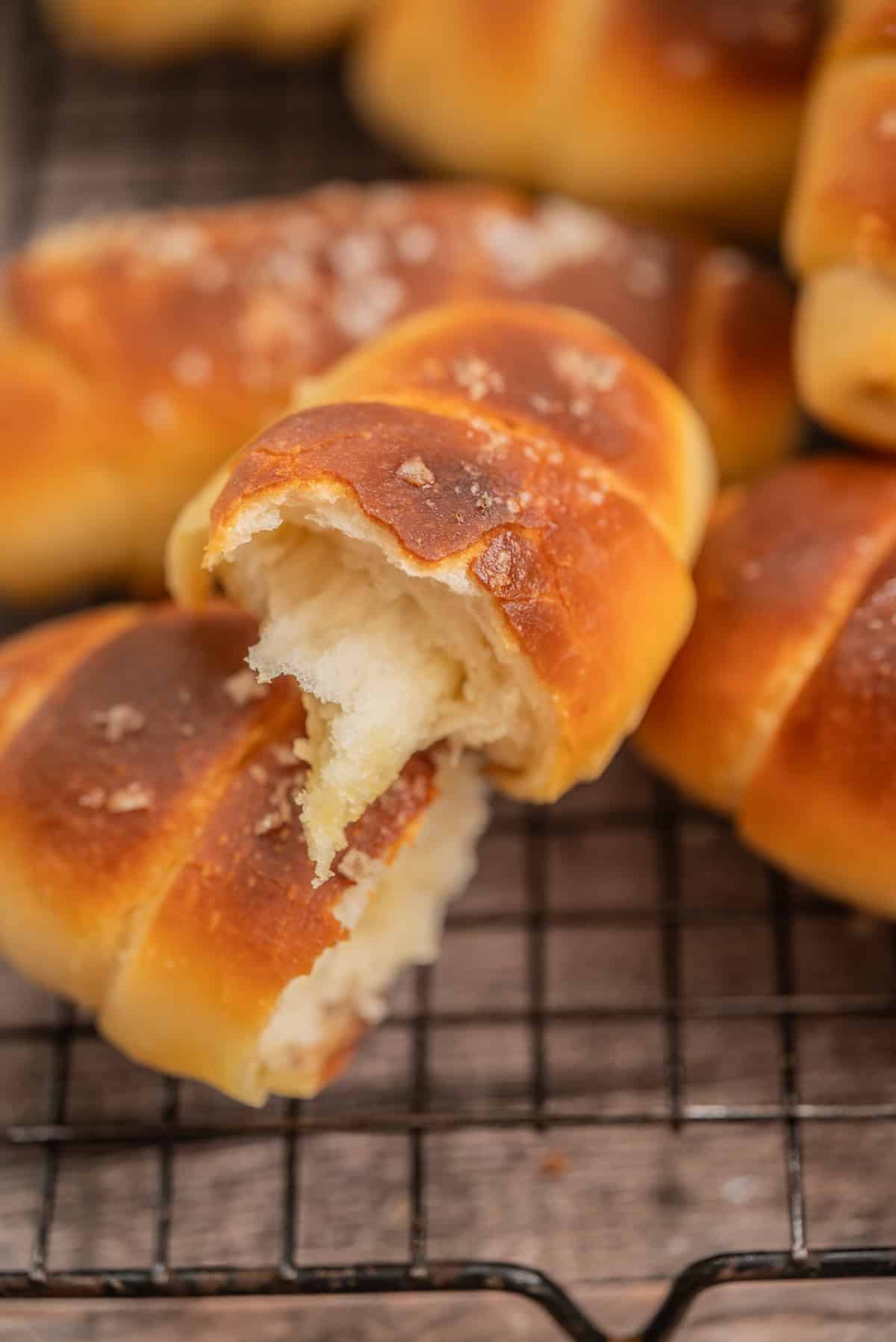 A close-up of a golden-brown, crescent-shaped bread roll with a soft, fluffy interior, partially torn open, resting on a cooling rack. Other similar rolls are visible in the background.
