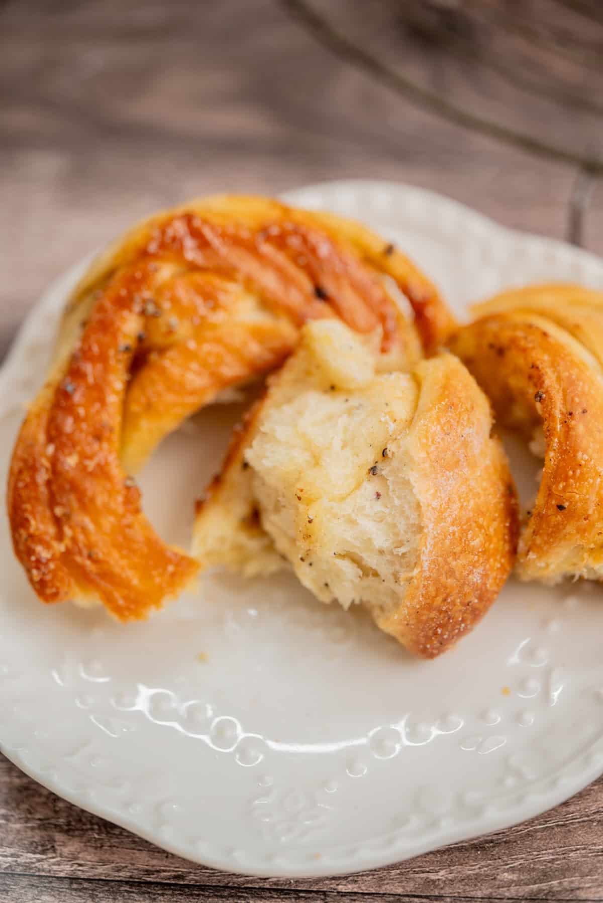 A close-up of braided pieces of golden, fluffy bread with a soft interior, served on a white decorative plate atop a wooden surface.