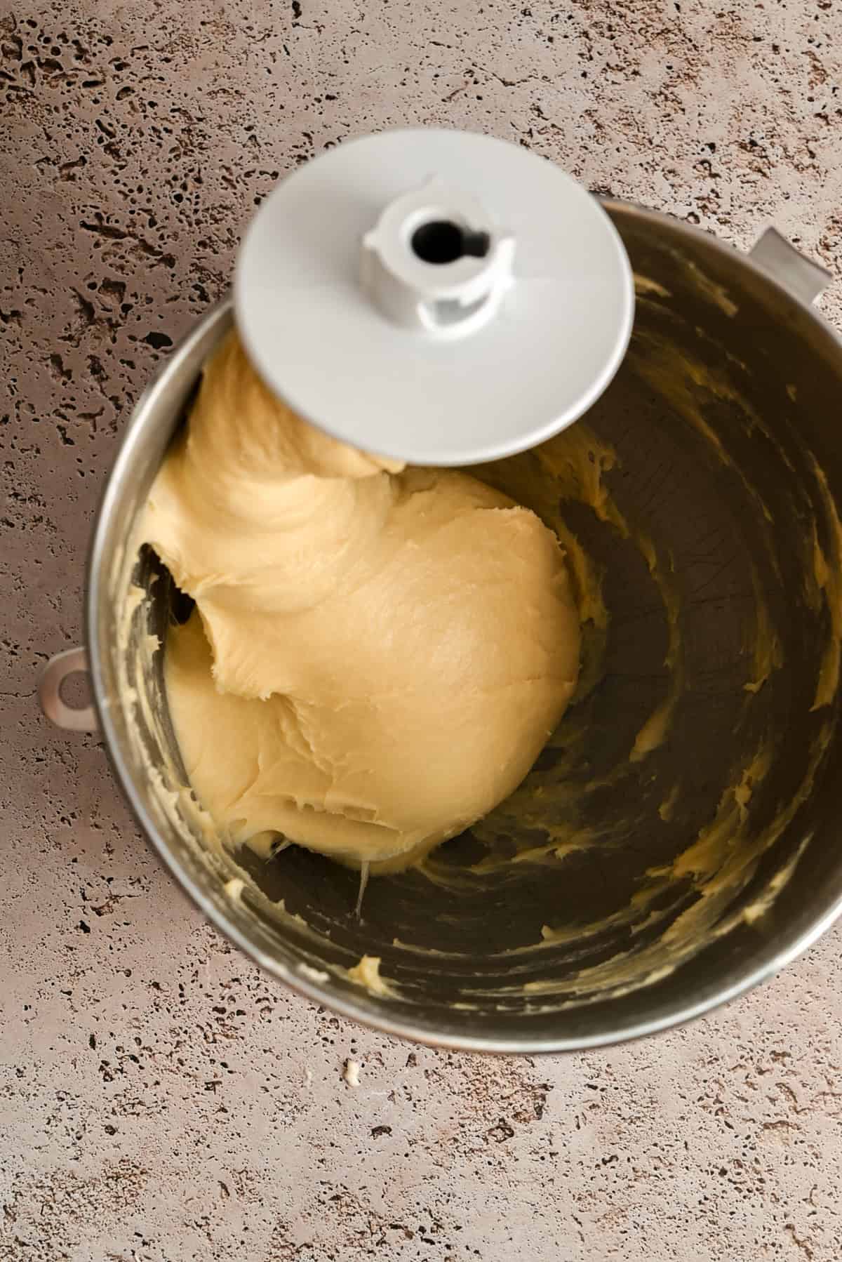 A stand mixer bowl filled with smooth, pale yellow dough being mixed by the dough hook attachment, set on a speckled beige countertop.