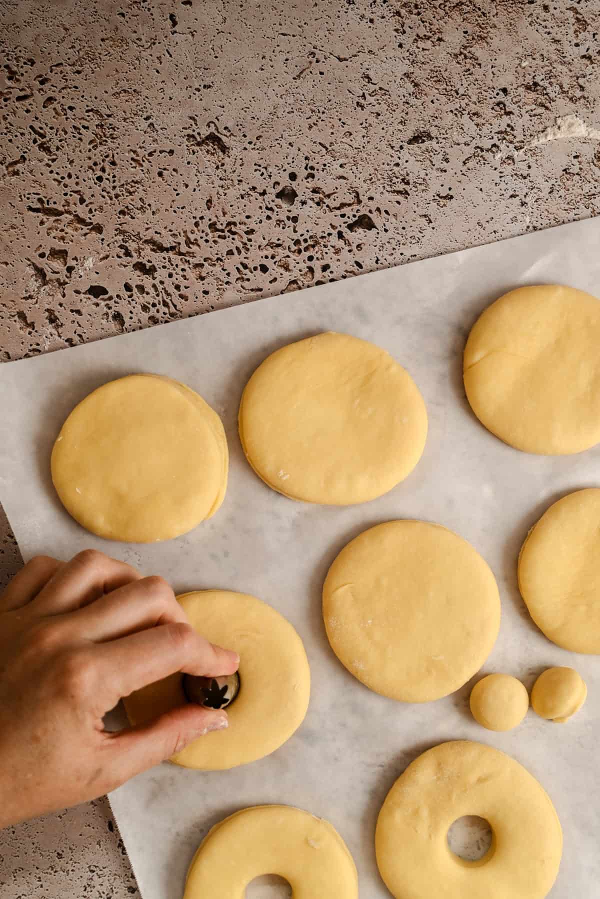 A hand presses a small round cutter into circular dough pieces on parchment paper, making shapes for baking. The countertop is speckled, and several dough circles are already cut.