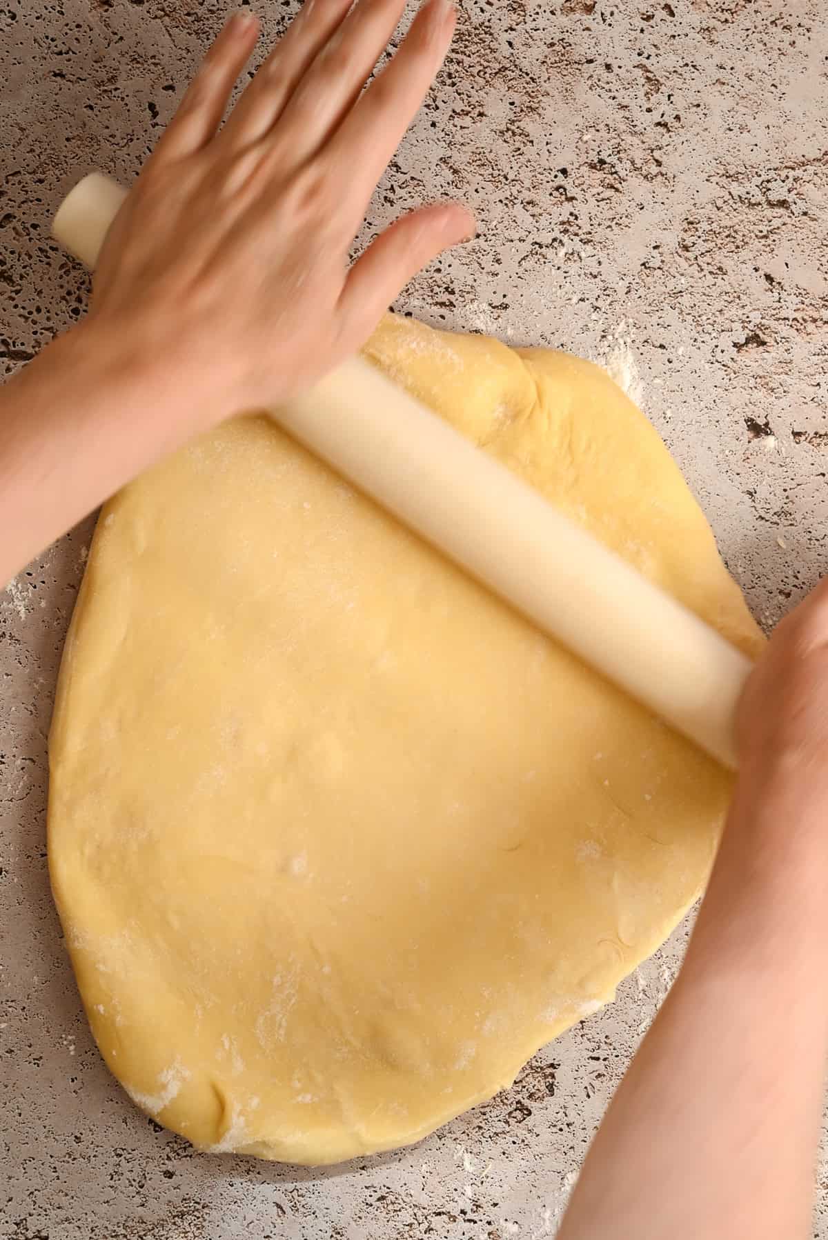 Two hands use a rolling pin to flatten a large piece of dough on a lightly floured surface. The dough is yellow and smooth, and the surface has a speckled, stone-like appearance.