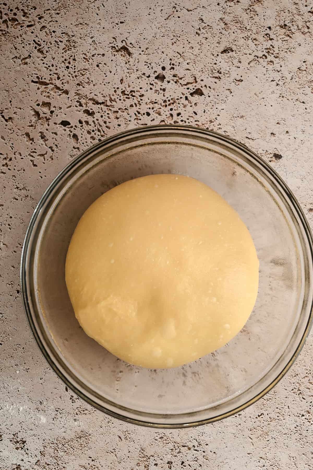 A round ball of risen dough sits in a clear glass bowl on a speckled countertop.
