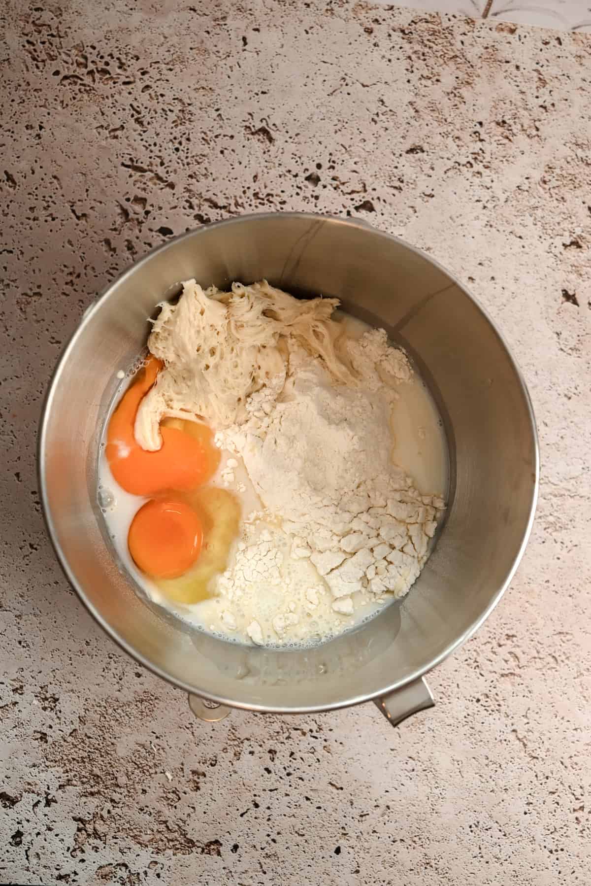 A metal mixing bowl contains two eggs, flour, dough, and milk, all set on a textured beige countertop. The ingredients are unmixed and ready for baking preparation.
