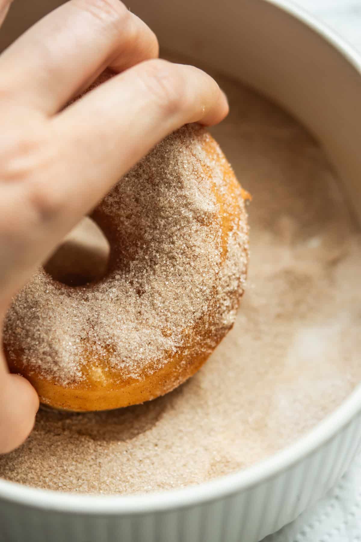 A hand dips a freshly baked donut into a bowl of cinnamon sugar, coating its surface. The donut is golden brown and the bowl is white with ridged sides.