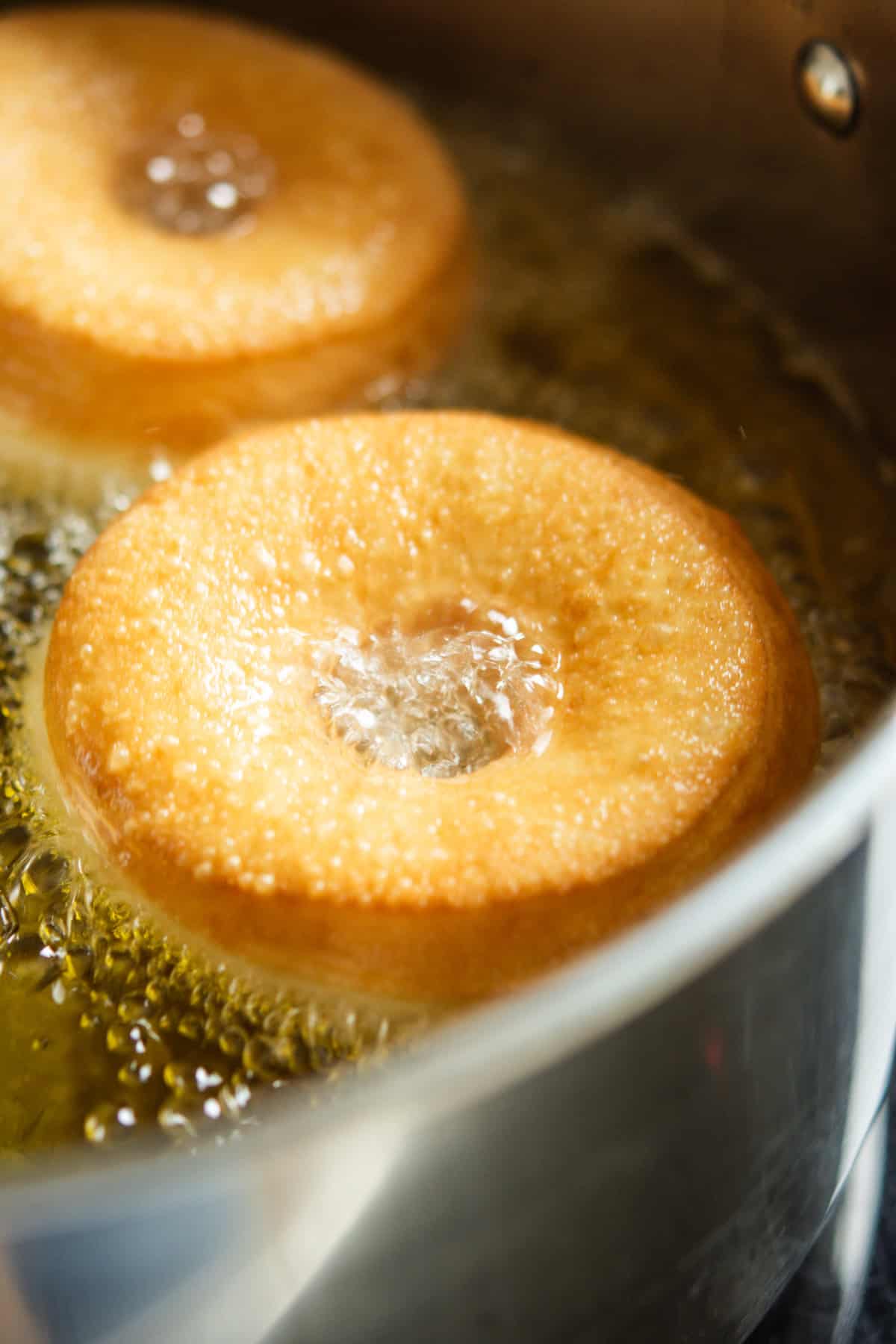Two doughnuts are being deep-fried in hot oil inside a metal pot, with bubbles forming around the edges of the doughnuts as they cook and turn golden brown.
