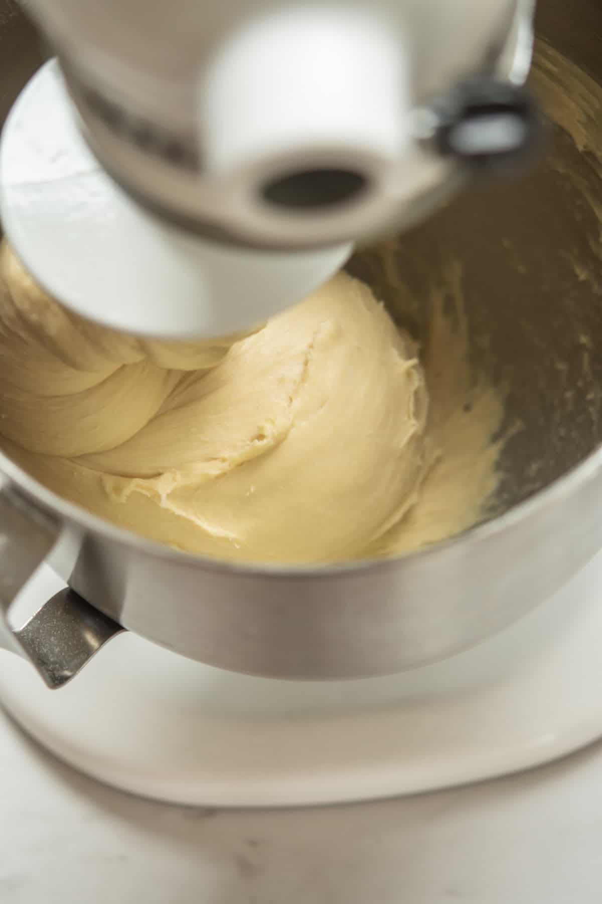 A close-up of a stand mixer blending dough in a stainless steel bowl, with the dough hook attachment kneading the smooth, creamy mixture.