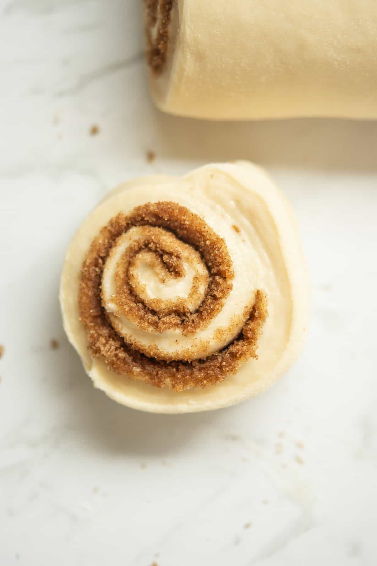 A close-up of an unbaked cinnamon roll with visible layers of dough and cinnamon sugar filling, resting on a white surface.