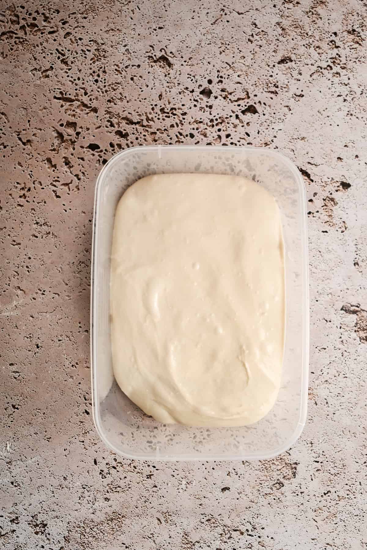 A rectangular plastic container filled with smooth, creamy dough sits on a textured, light brown surface.
