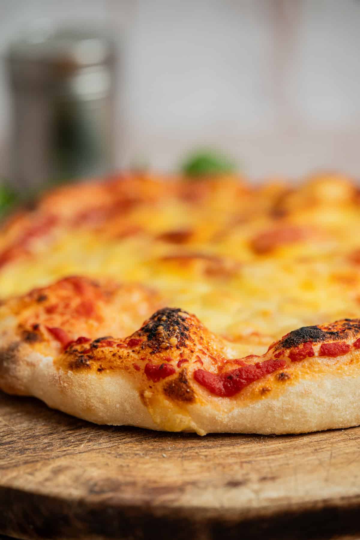 A close-up of a freshly baked pizza with a puffy, golden-brown crust, melted cheese, and tomato sauce, sitting on a wooden surface. The pizza looks rustic with slightly charred edges.
