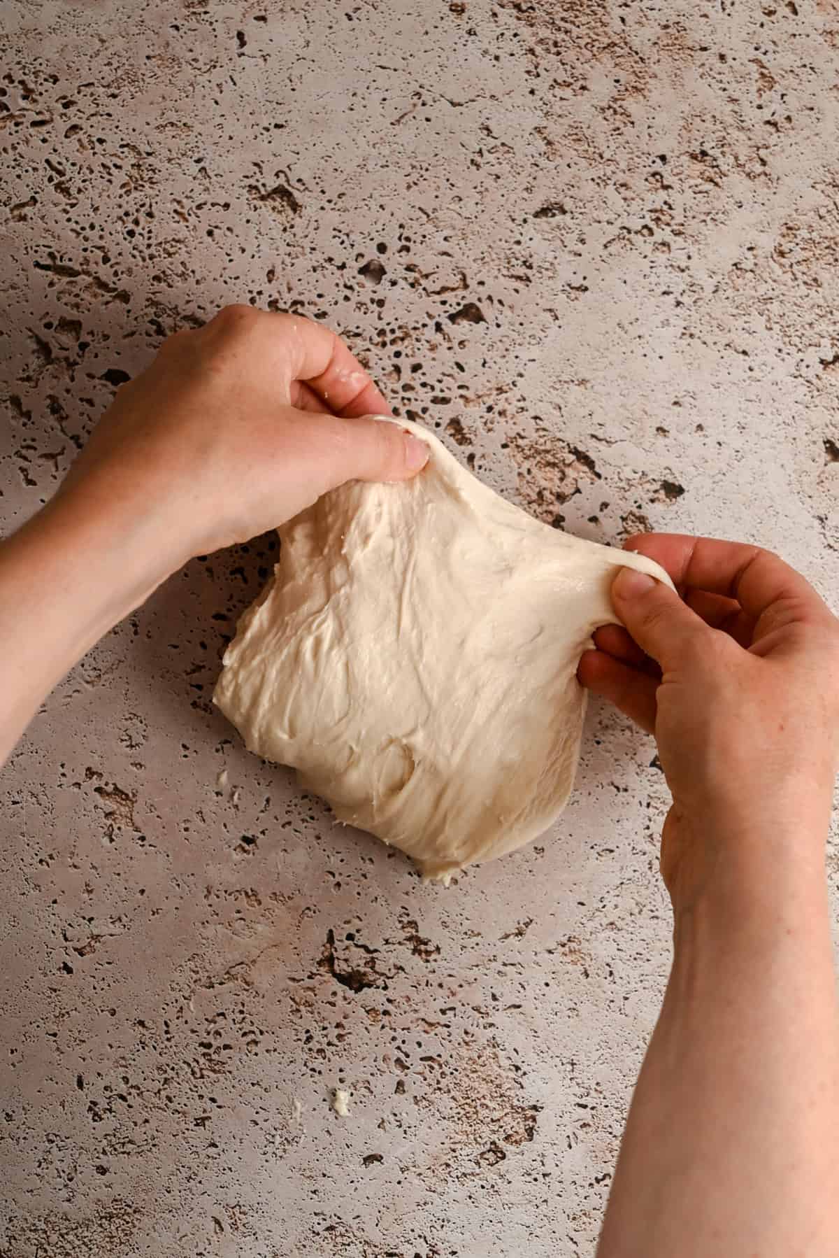 Two hands stretch a piece of dough on a textured, light-colored surface, demonstrating a baking or cooking process.