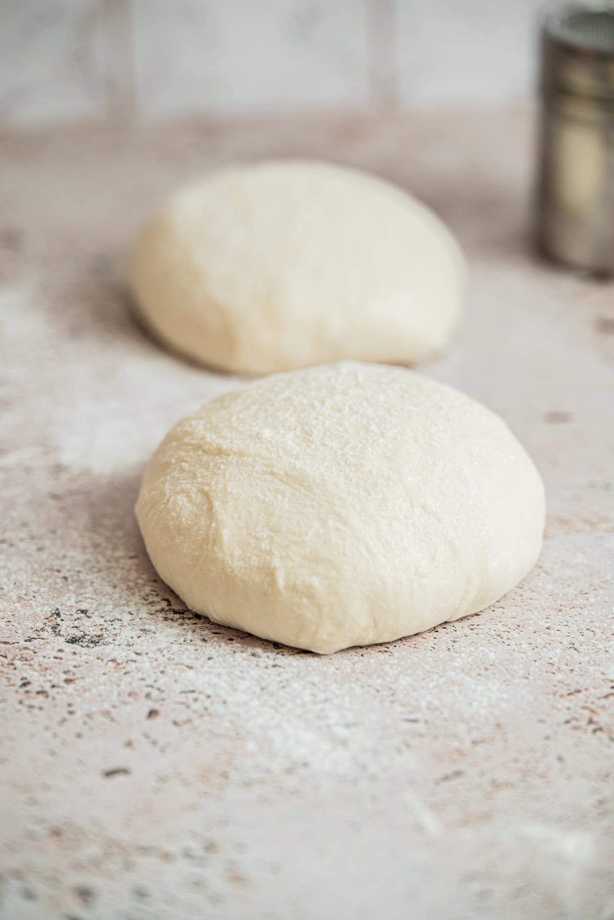 Two round balls of dough rest on a floured surface, ready for baking. A metal container, likely a flour shaker, is visible in the background. The scene suggests bread or pizza preparation.