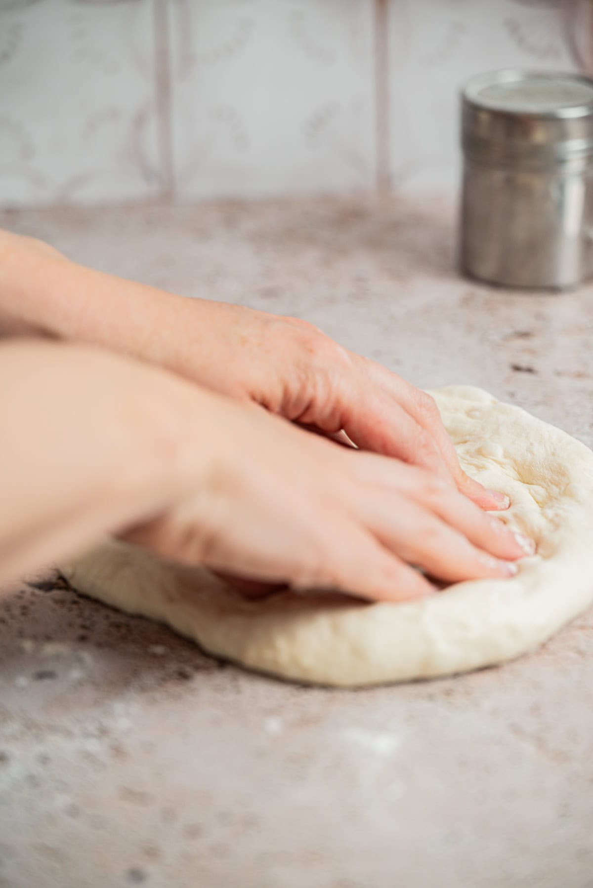 Two hands pressing and shaping pizza dough on a lightly floured countertop, with a metal flour shaker in the background.