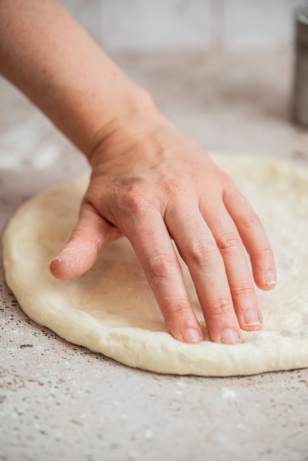 A close-up of a hand pressing and shaping raw pizza dough on a floured surface.