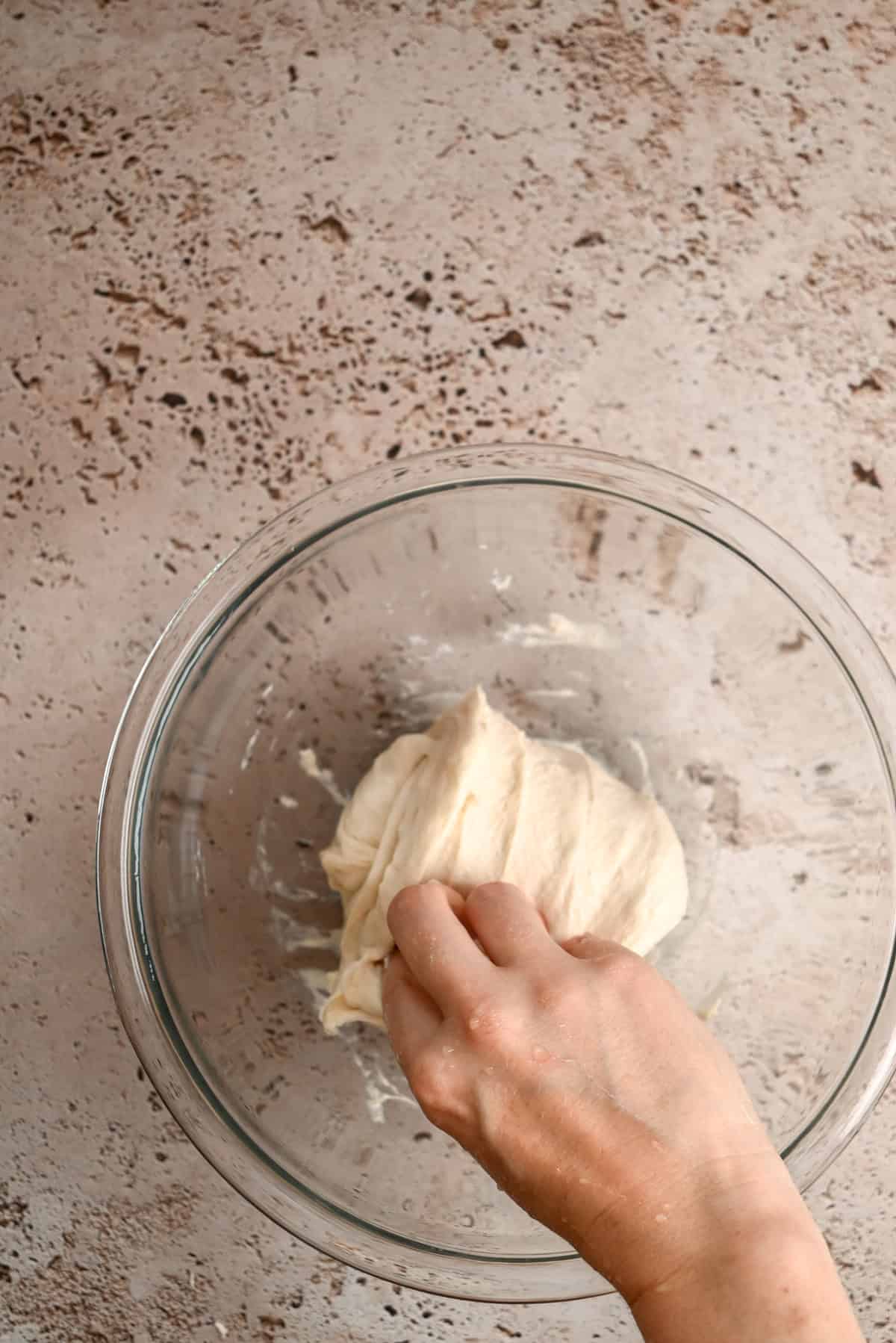 A hand kneads a piece of dough in a clear glass bowl on a speckled, light-colored countertop.