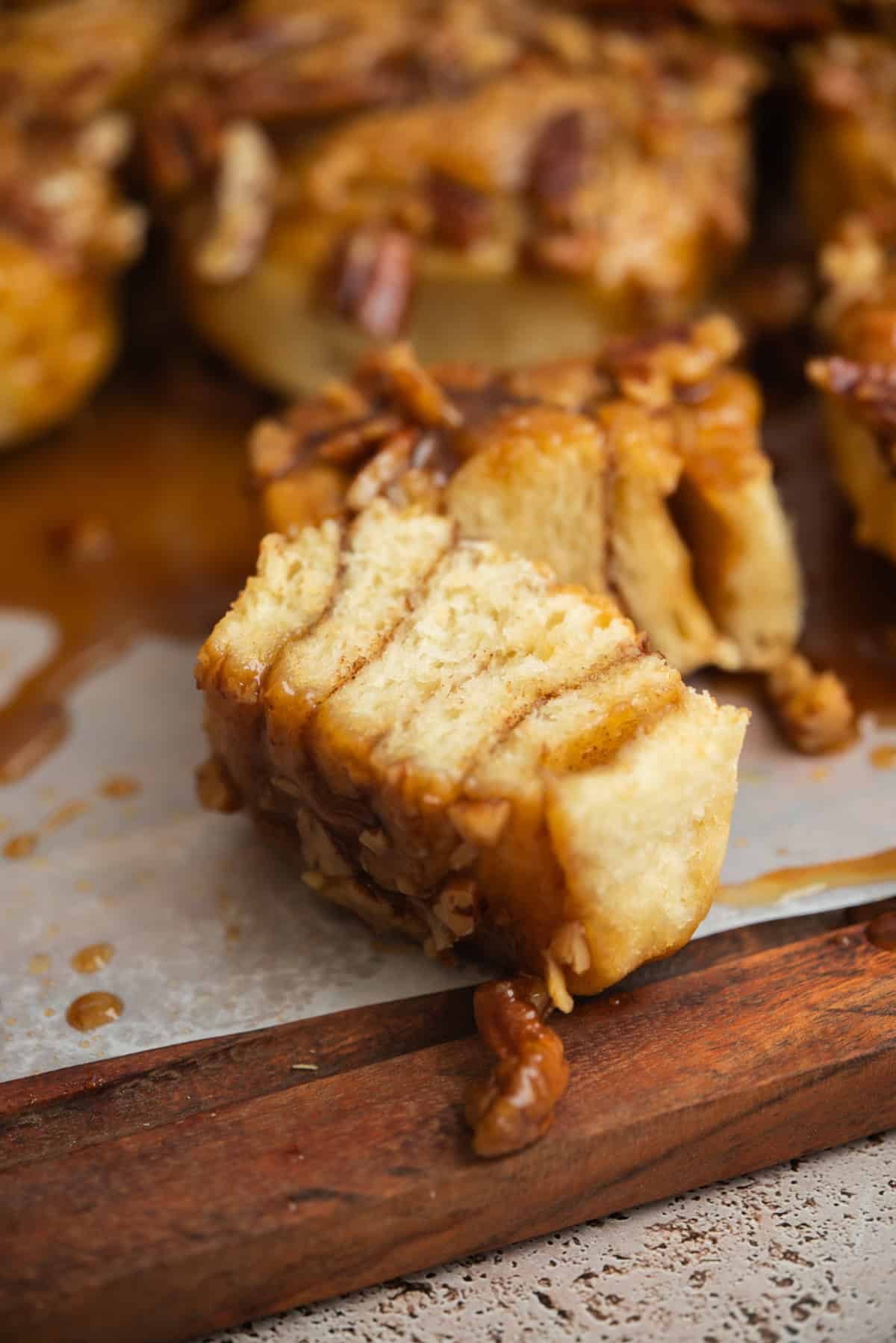 A close-up of a sticky sourdough pecan cinnamon roll, partially pulled apart to show its soft, layered texture and caramelized topping, resting on parchment paper with pecans scattered around.
