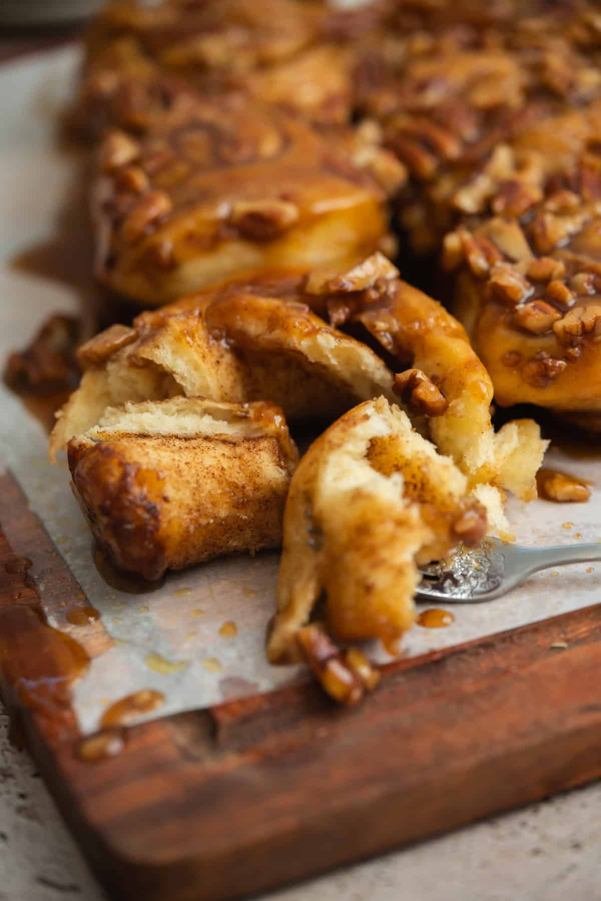 Close-up of sticky sourdough sticky buns on parchment paper, with one roll pulled apart by a fork, showing the soft, fluffy interior and caramel glaze with chopped pecans on top.