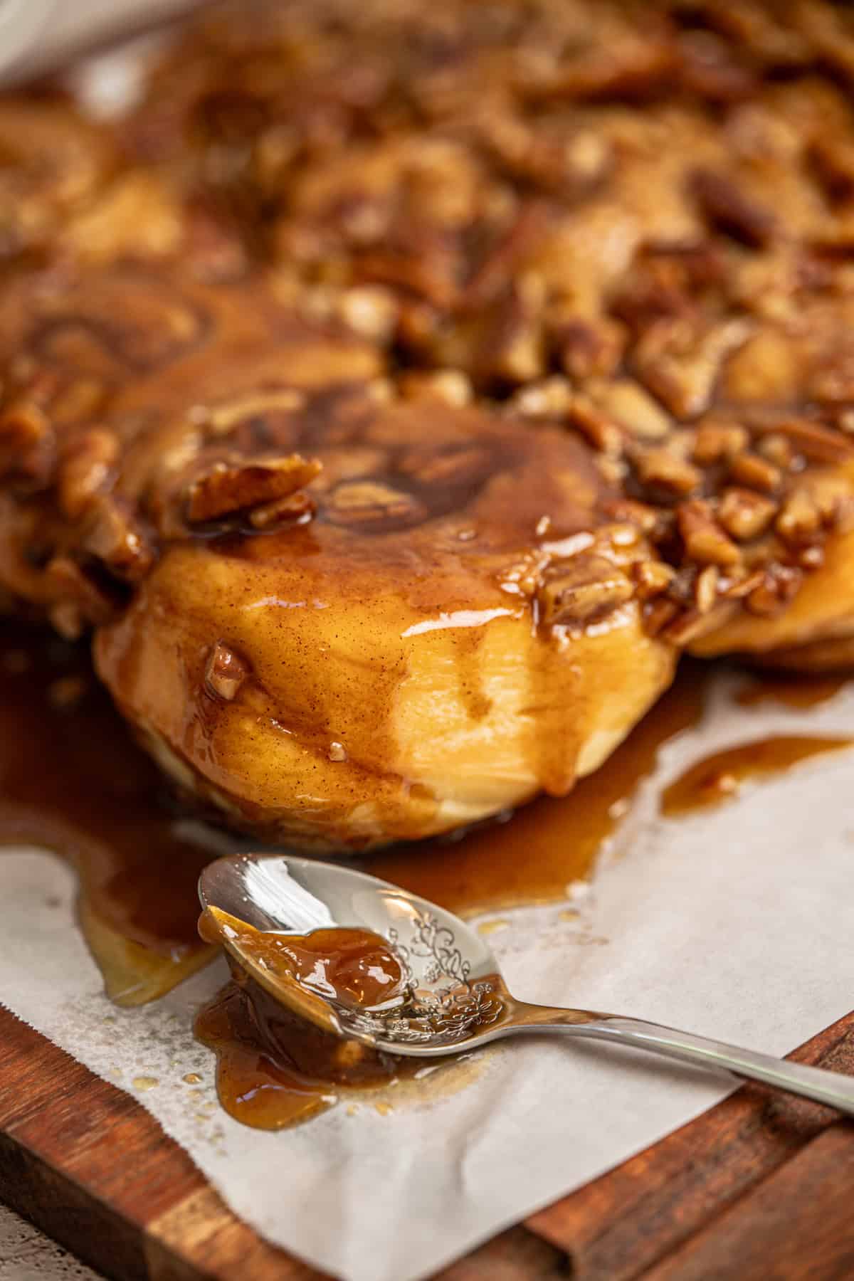 Close-up of sourdough sticky buns topped with chopped pecans and caramel glaze on parchment paper, with a spoon covered in glaze resting in the foreground.