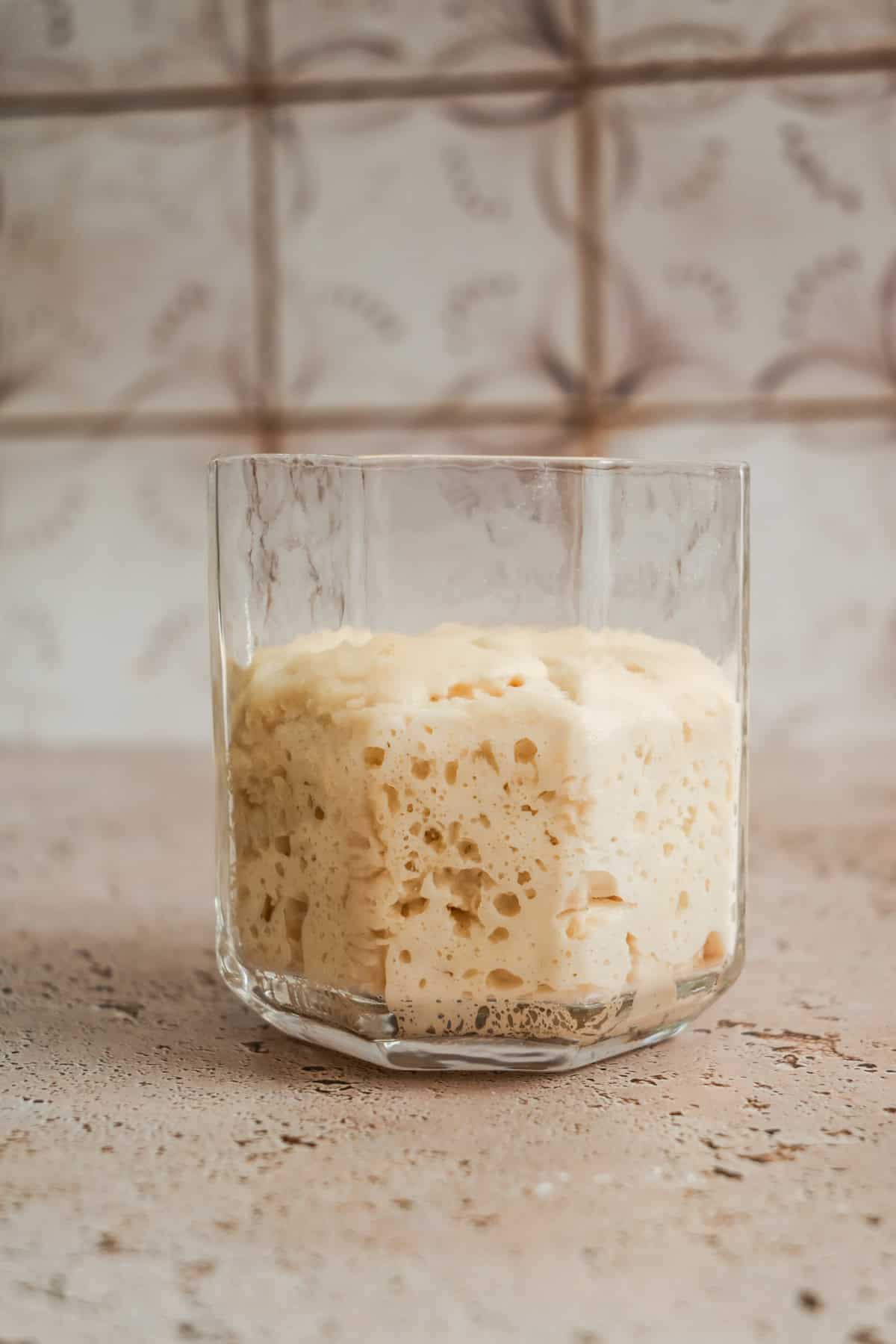 A glass cup contains a single serving of light, fluffy bread with visible air holes, resembling mug bread or microwave sponge bread, set on a textured countertop with a patterned tile background.
