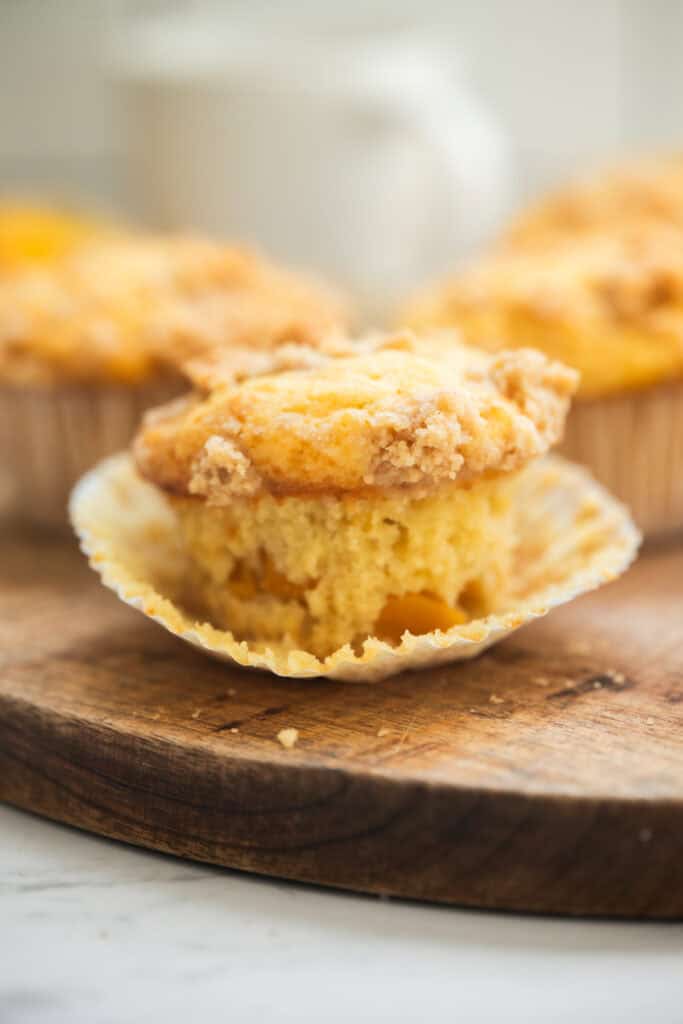 A close-up of a crumb-topped muffin with a partially peeled paper liner, sitting on a wooden surface. Other muffins are blurred in the background. The muffin looks moist and fluffy inside.