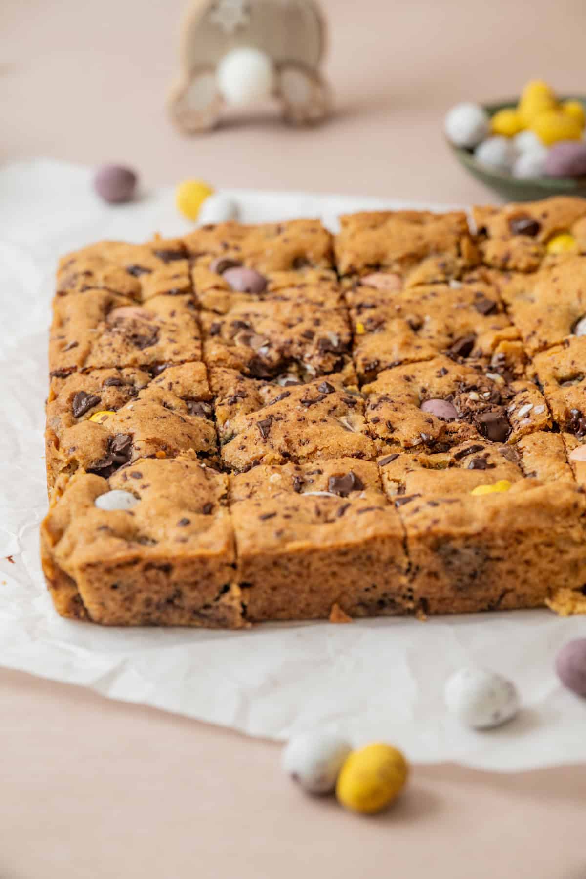 A square traybake of cookie bars, topped with chopped chocolate and colorful chocolate eggs, cut into squares and placed on white parchment paper. Scattered mini chocolate eggs are visible around the traybake.
