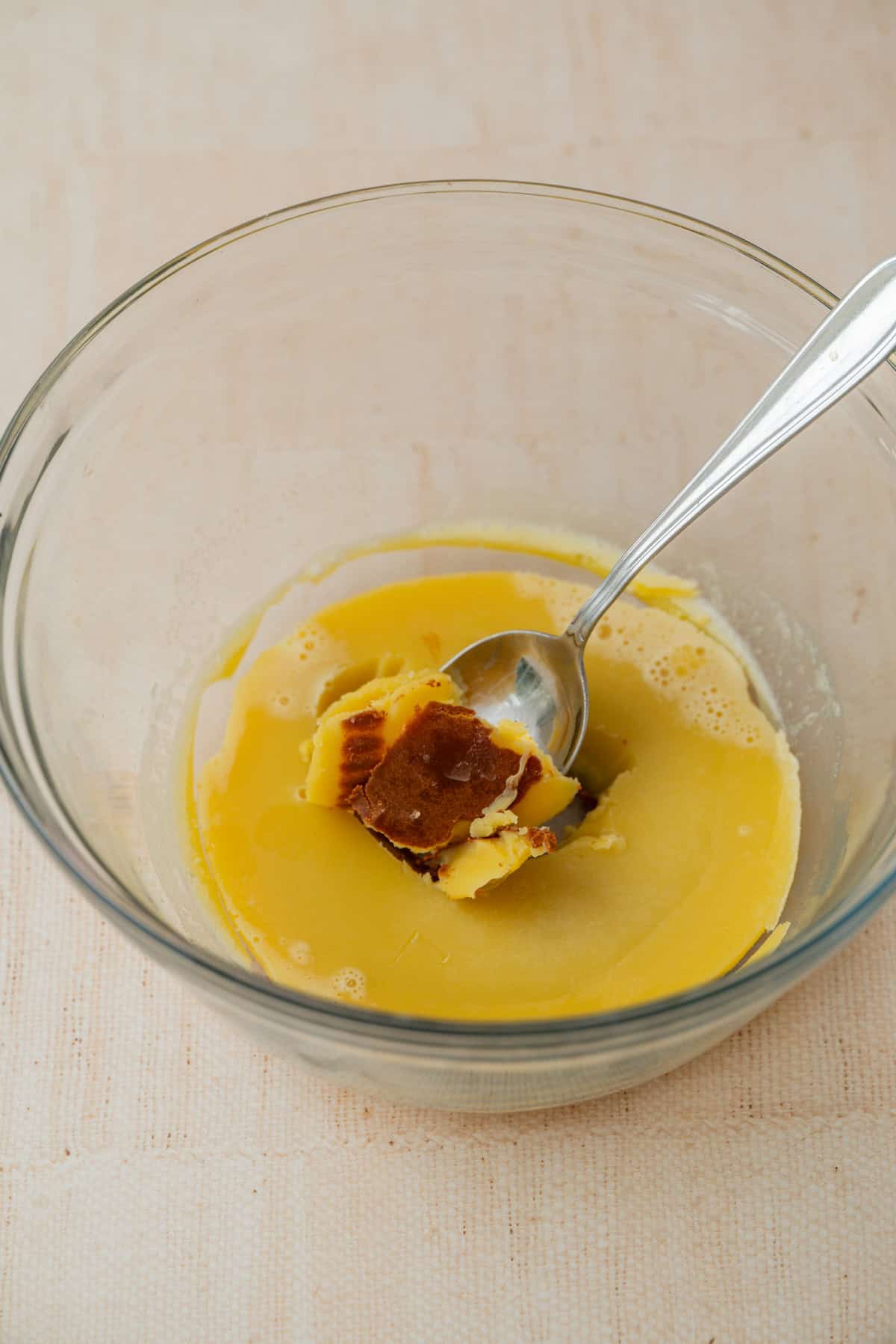 A spoon rests in a glass bowl containing solid and melted brown and yellow butter, likely browned butter, on a light-colored surface.