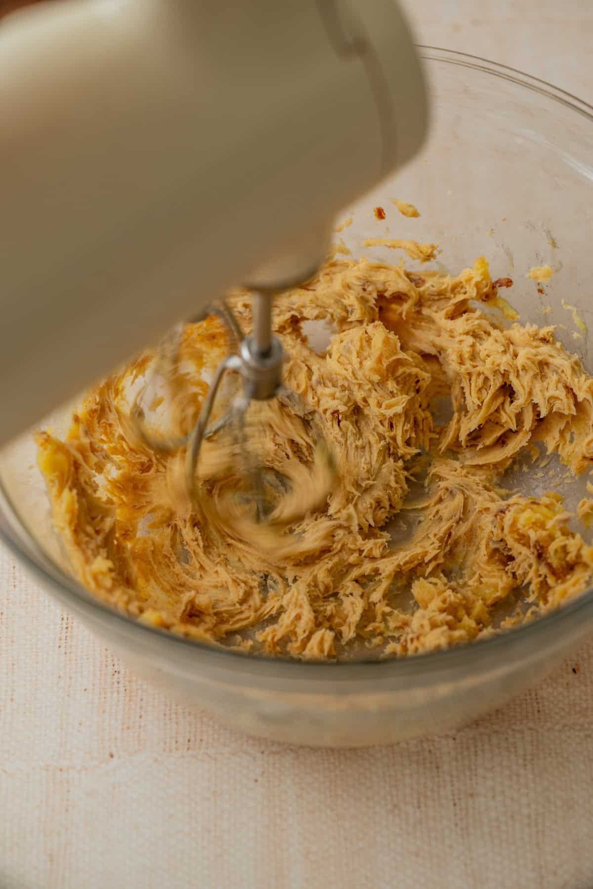 A close-up of an electric hand mixer blending a brown butter in a clear glass bowl on a light-colored surface.