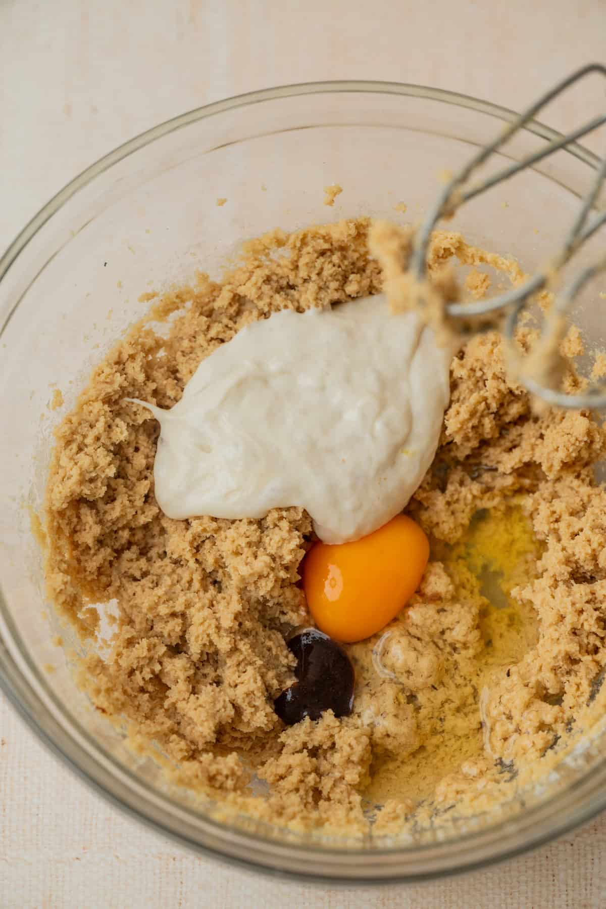 A glass bowl with cookie dough ingredients: creamed butter and sugar, a cracked egg, vanilla extract, and mashed banana, shown before being mixed together. An electric mixer beater is visible in the corner.