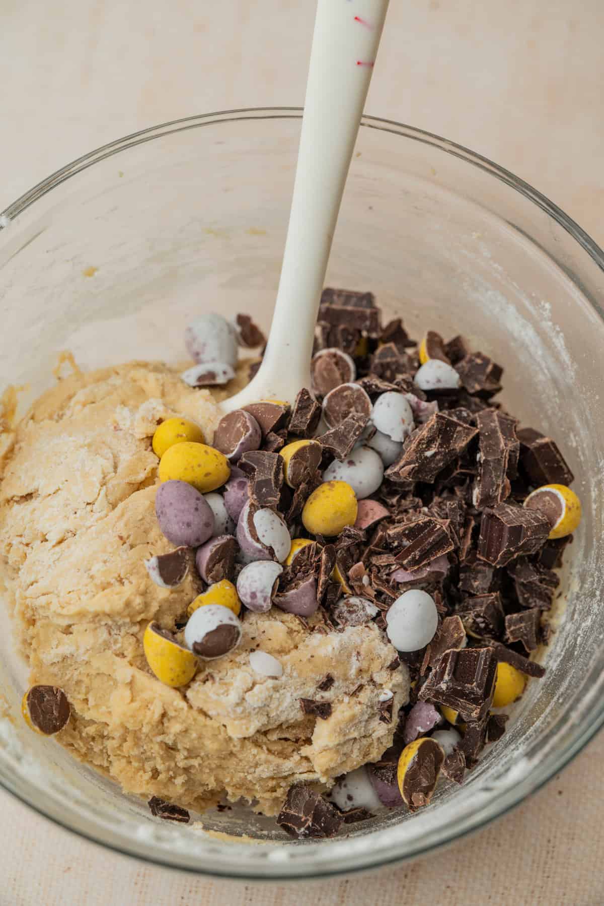 A glass bowl contains cookie dough mixed with chocolate chunks and colorful candy-coated chocolate eggs. A white spatula is partially submerged in the mixture.