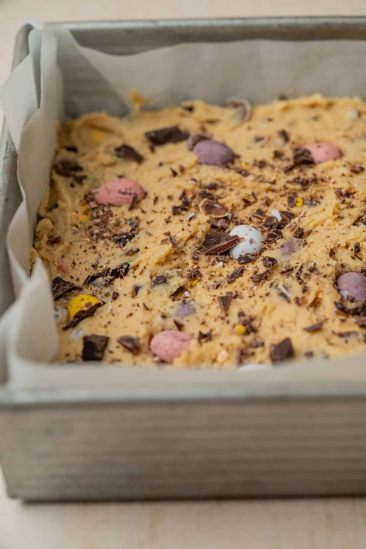 A square baking pan lined with parchment paper, filled with unbaked cookie dough mixed with chocolate chunks and colorful candy-coated chocolate eggs.