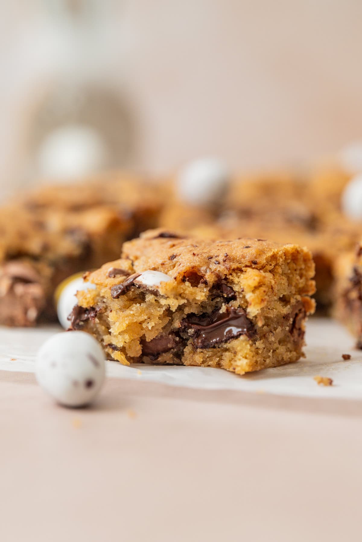 A close-up of a chocolate chip blondie with a gooey melted chocolate center, surrounded by blurred mini chocolate eggs and more blondies in the background.