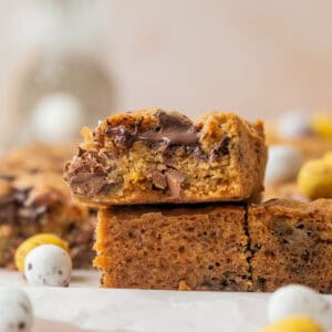 A close-up of thick, golden-brown Sourdough Mini Egg Cookie Bars with chocolate chunks, stacked on parchment paper. Colorful mini chocolate eggs are scattered around the bars, creating a festive, spring-themed scene.