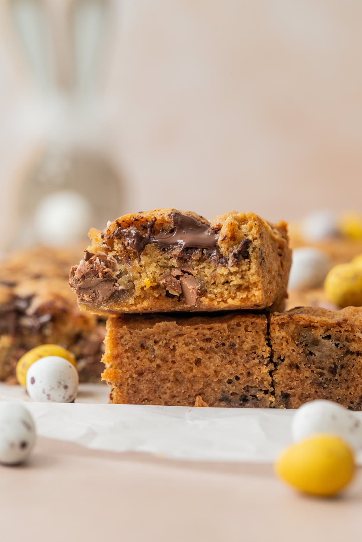 A close-up of thick, golden-brown Sourdough Mini Egg Cookie Bars with chocolate chunks, stacked on parchment paper. Colorful mini chocolate eggs are scattered around the bars, creating a festive, spring-themed scene.