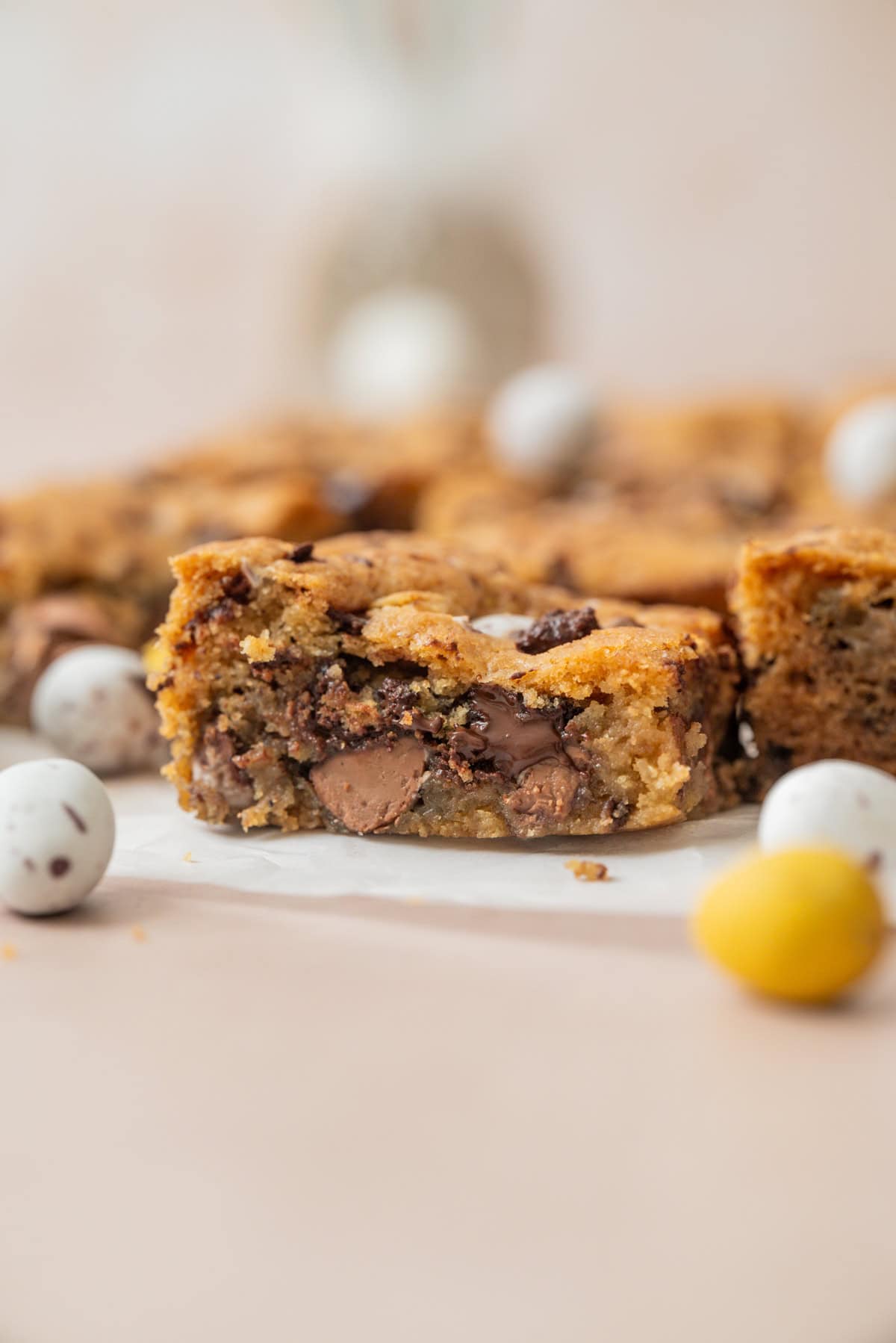 A close-up of a thick, gooey chocolate chip cookie bar with chunks of chocolate and mini chocolate eggs inside. More cookie bars and chocolate eggs are blurred in the background.