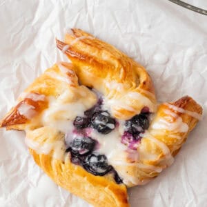 A golden, flaky pastry topped with blueberries and drizzled with white icing sits on crinkled parchment paper.