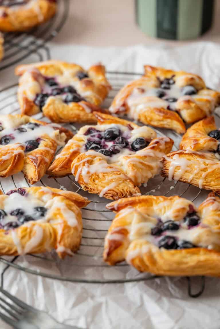 Freshly baked puff pastries topped with blueberries and drizzled with white icing are arranged on a round cooling rack, with a crinkled white paper beneath them.