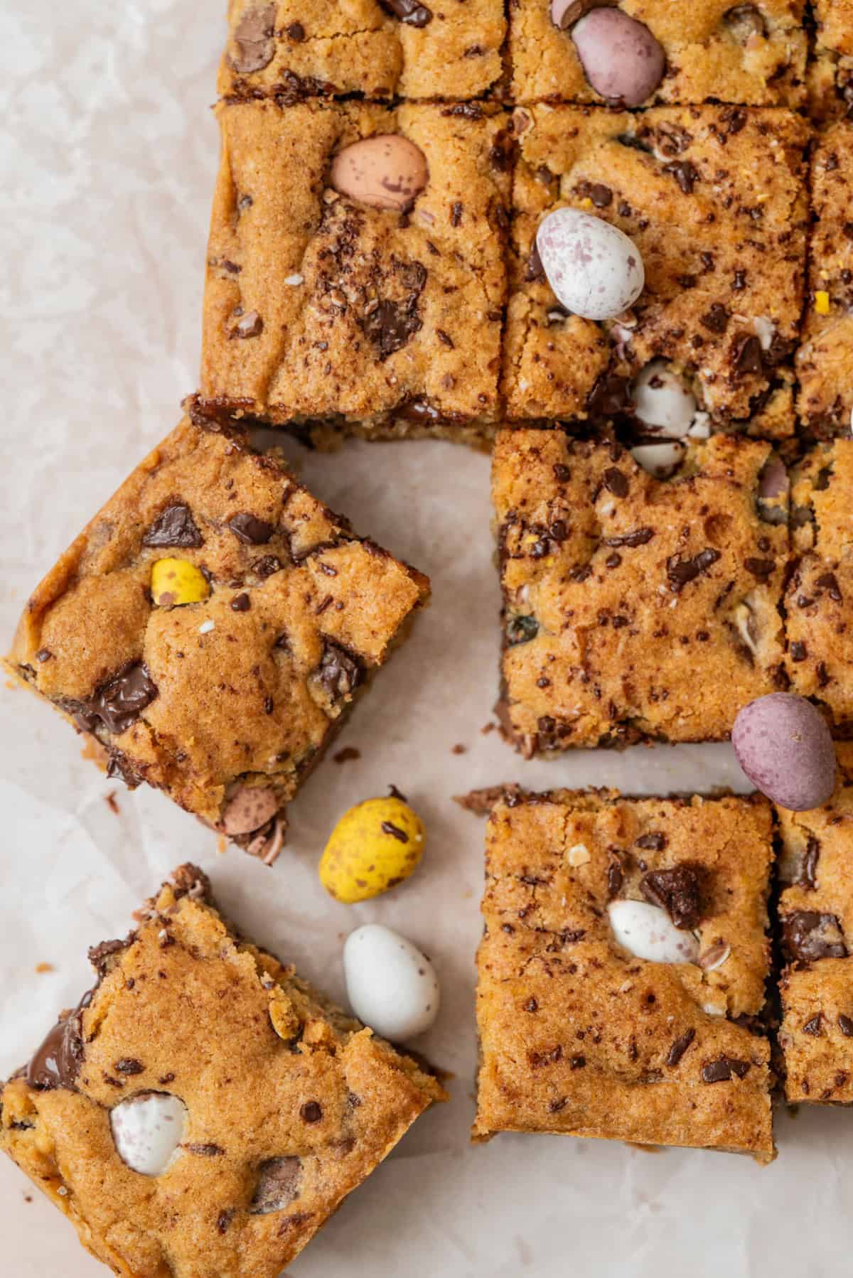 Top-down view of sourdough mini egg cookie bars cut into squares, with colorful candy-coated chocolate eggs scattered on and within the bars, set on a light marble surface.