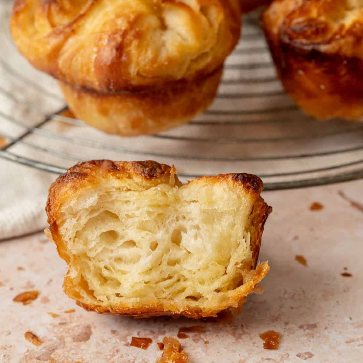 Close-up of a flaky, golden-brown pastry with a bite taken out, showing airy, layered texture inside. More pastries are blurred in the background on a round cooling rack.