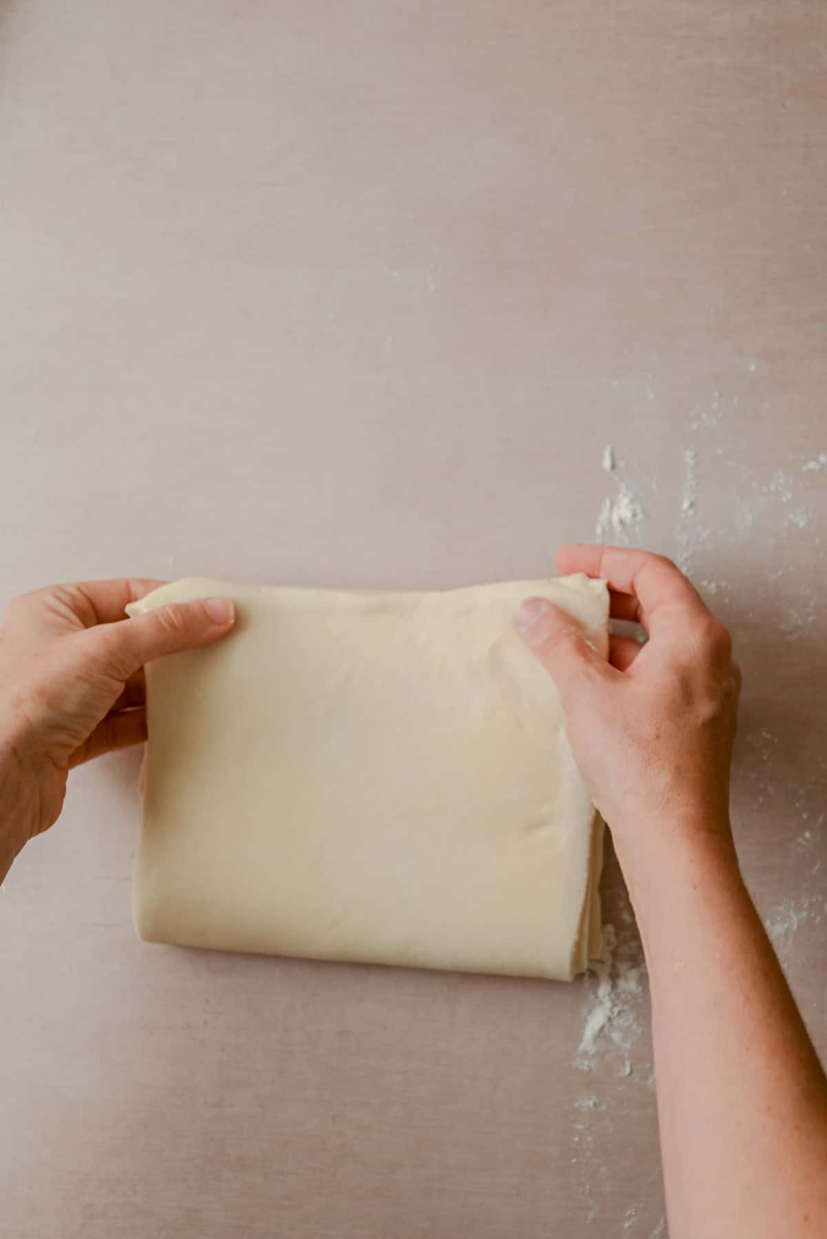 Hands folding a rectangular sheet of dough on a lightly floured surface.