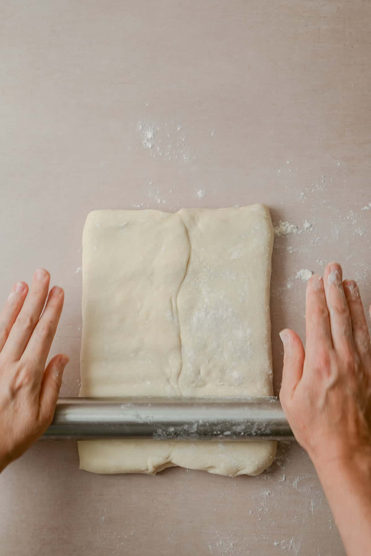 Two hands use a rolling pin to roll out a rectangular piece of dough on a lightly floured surface.