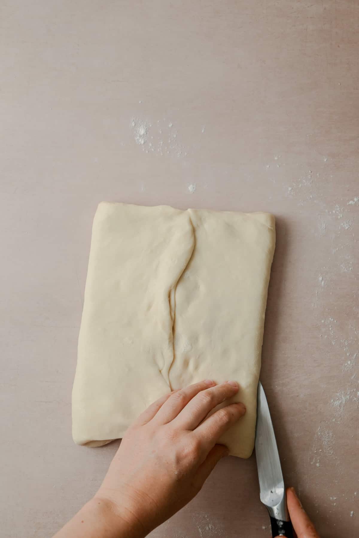 A person uses a knife to cut folded dough on a lightly floured surface, preparing it for baking.