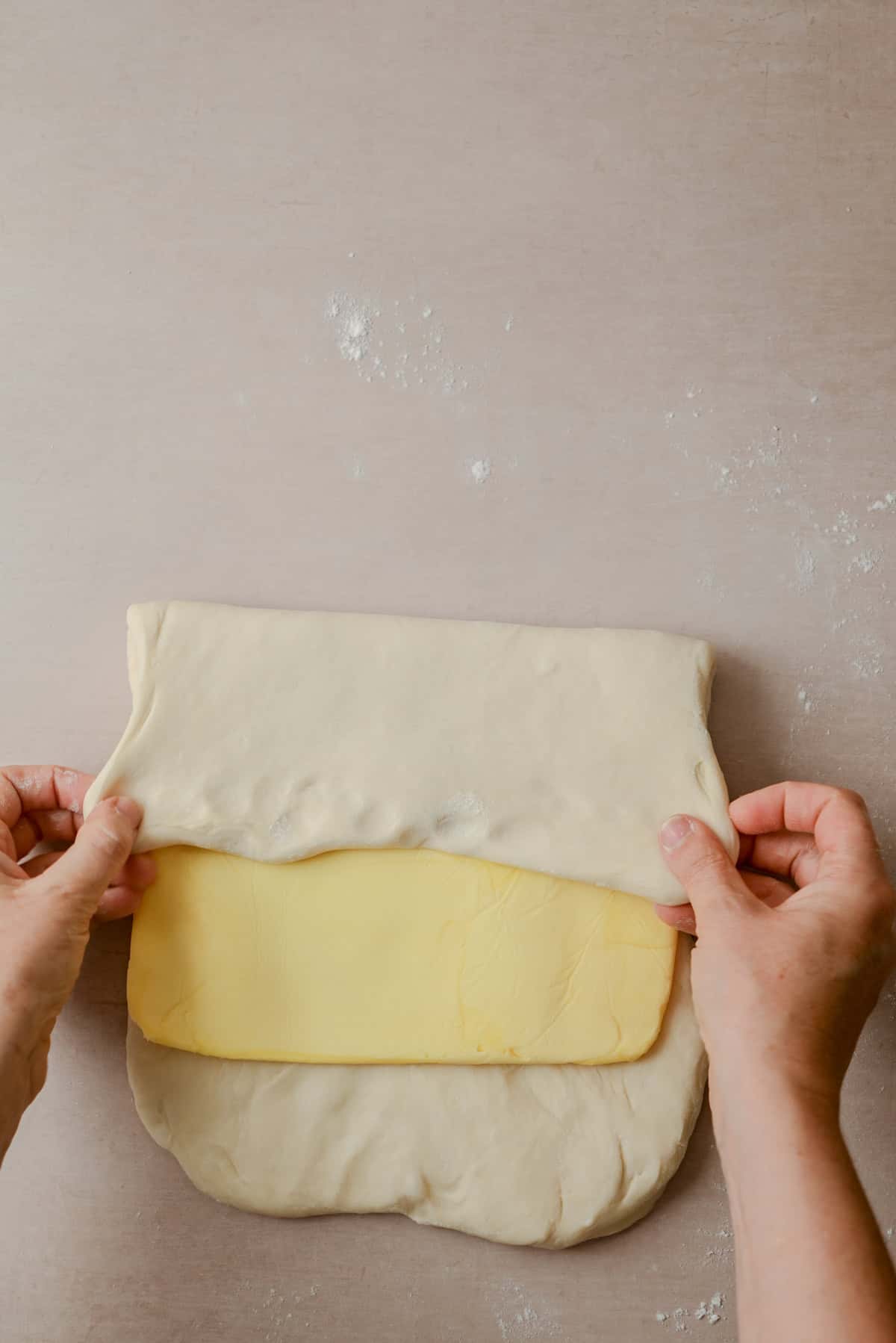 Hands fold a sheet of dough over a large rectangular slab of butter on a floured surface, preparing the dough for lamination, likely for making pastries such as croissants or puff pastry.