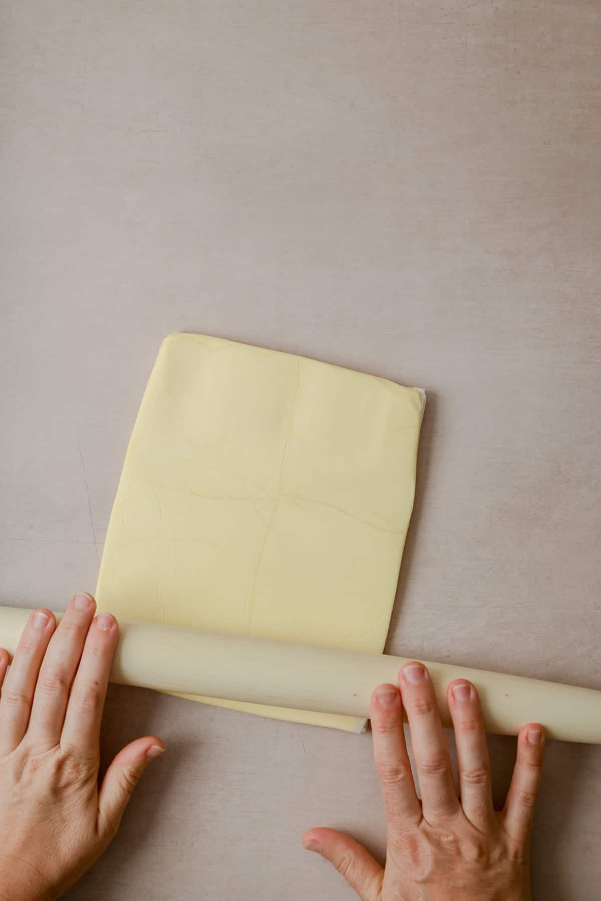 Two hands use a rolling pin to flatten a square sheet of dough on a light-colored surface.