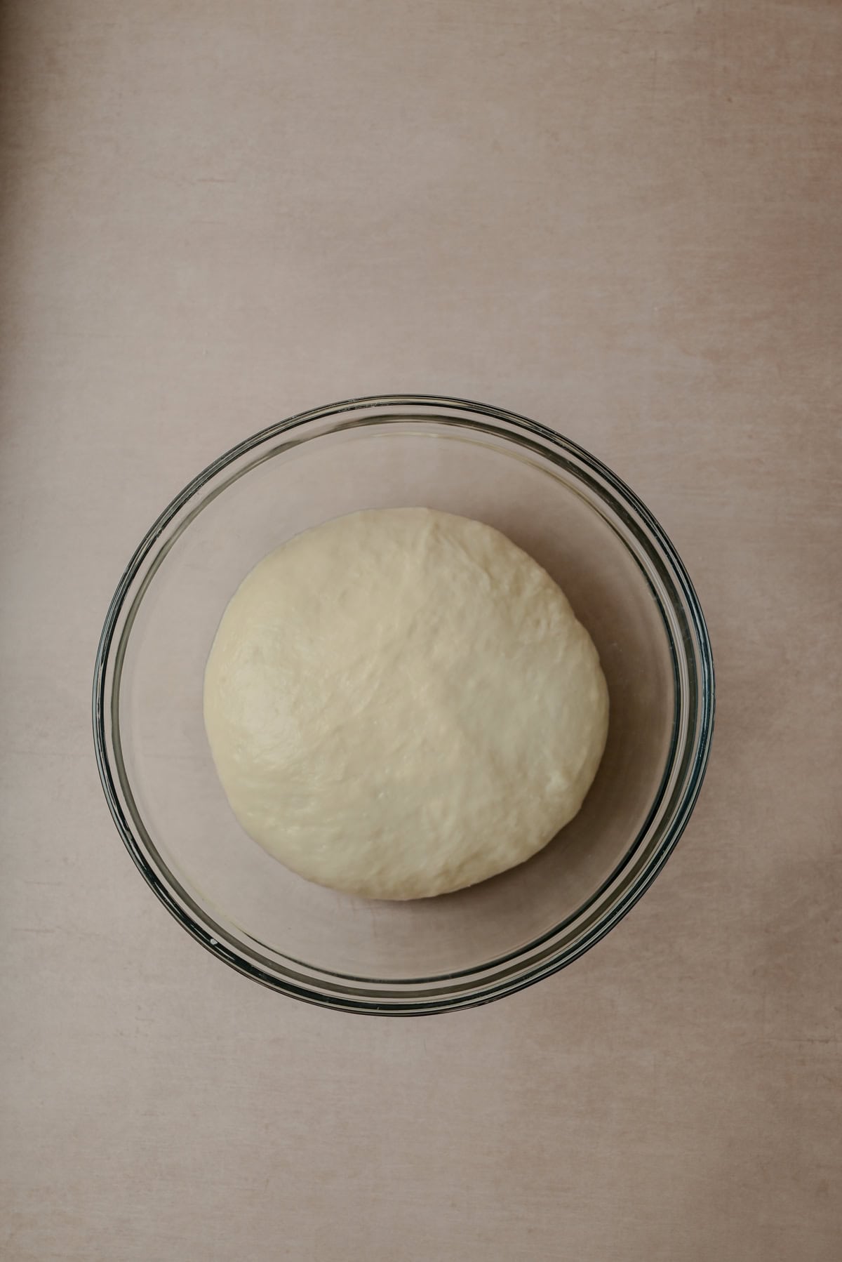A round ball of dough rests in a clear glass bowl on a light beige surface, ready to rise.