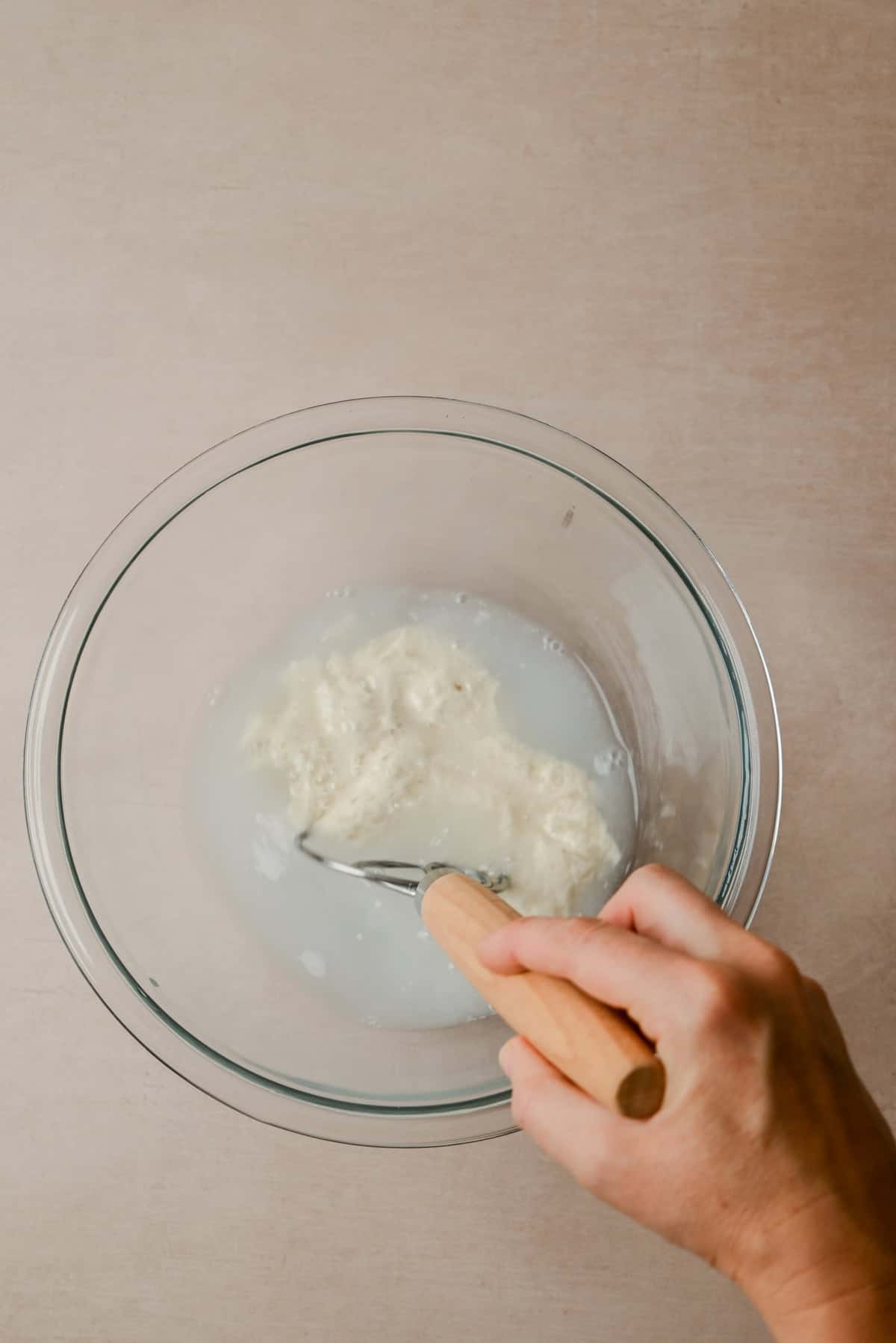 A hand holding a wooden-handled whisk is mixing a white, lumpy mixture in a clear glass bowl on a beige surface.