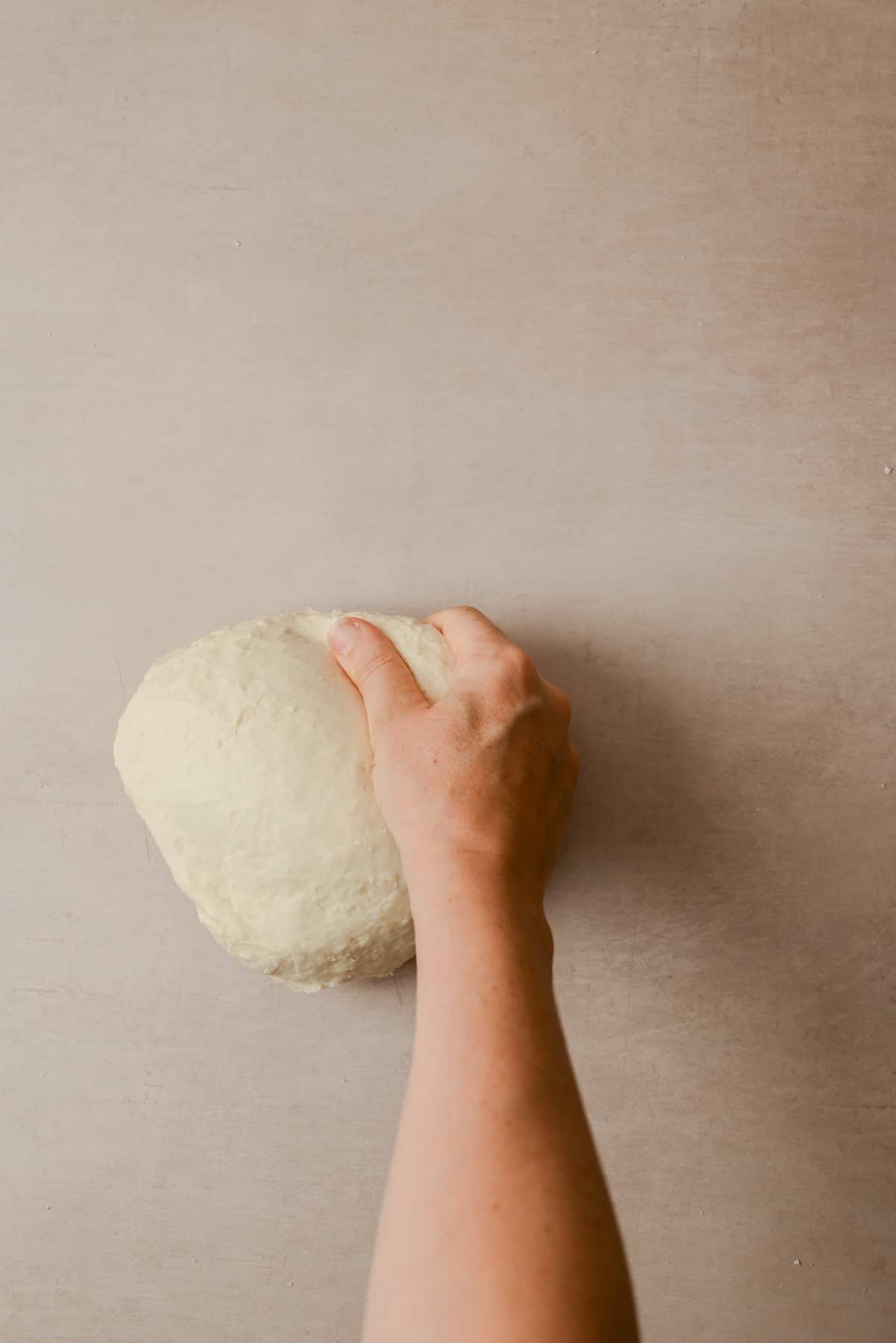 A hand kneads a ball of dough on a light brown surface.