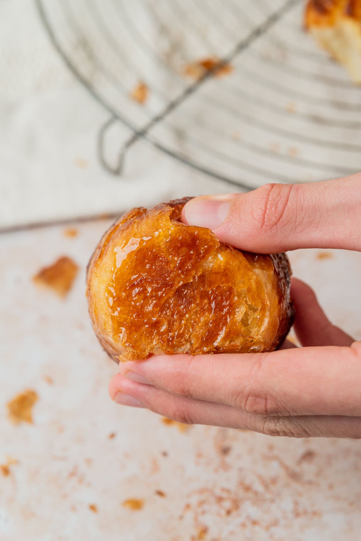 A hand holds a round, golden-brown pastry with a glossy, caramelized top. In the background, there is a wire cooling rack and scattered pastry crumbs on a light surface.