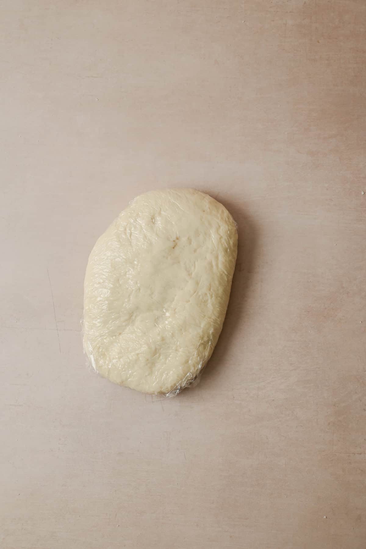 A ball of dough wrapped in plastic wrap rests on a light brown surface.