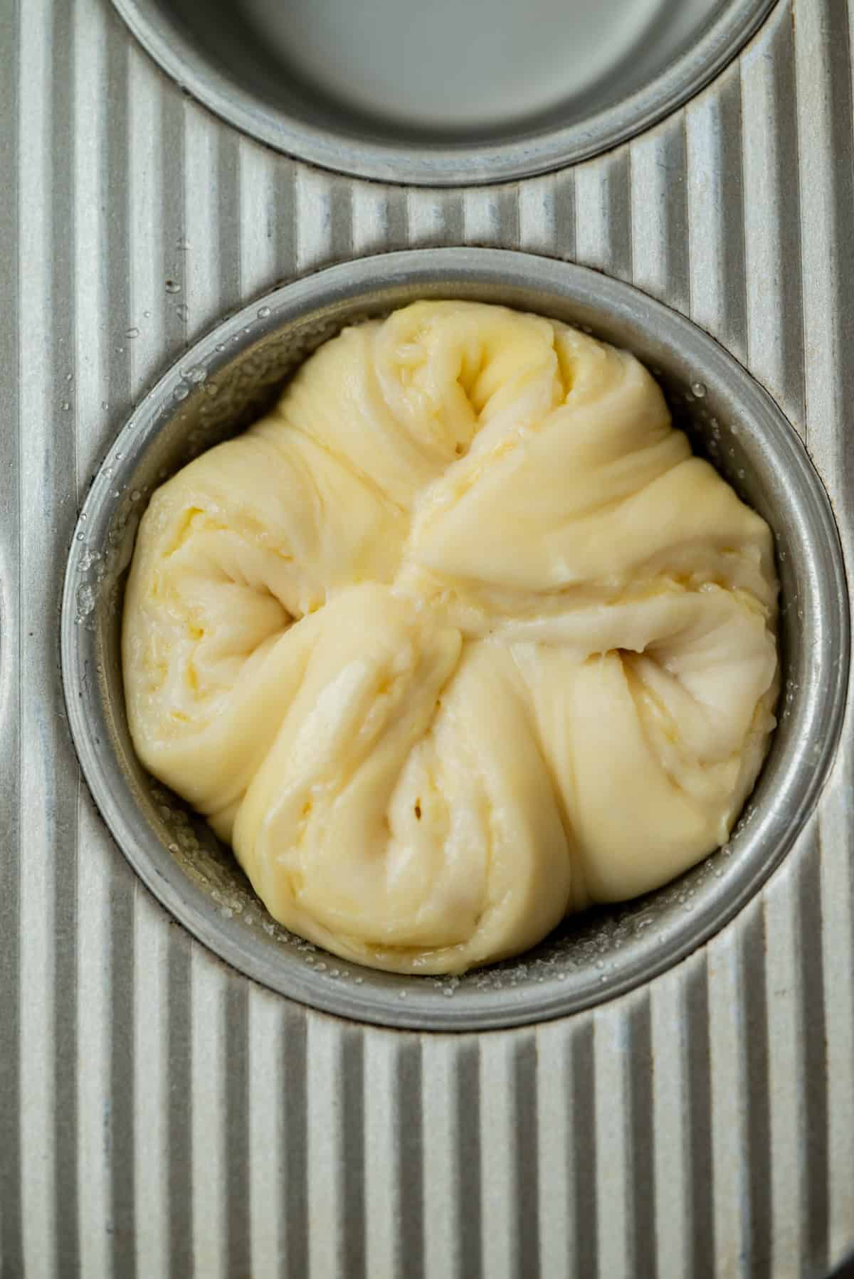Close-up of a muffin tin holding a single portion of unbaked, twisted dough with visible layers, ready to be baked. The metal tin has a ribbed texture and another empty mold is visible above.