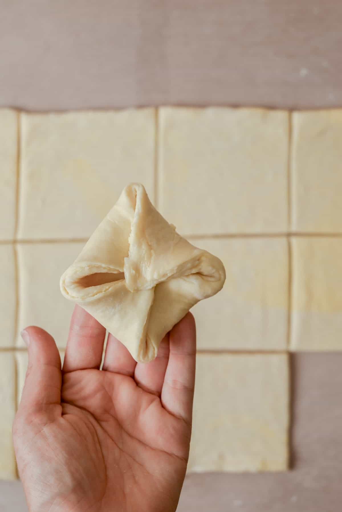 A hand holds a folded piece of dough shaped like a pouch, with several flat dough squares laid out on the surface in the background.